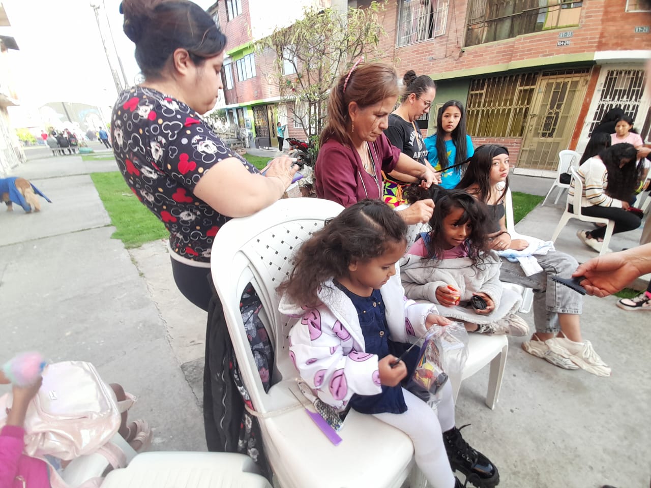 Voluntarias peinando a niñas sentadas en sillas plásticas en la calle, Fundación Rosas del Carmelo, Bogotá