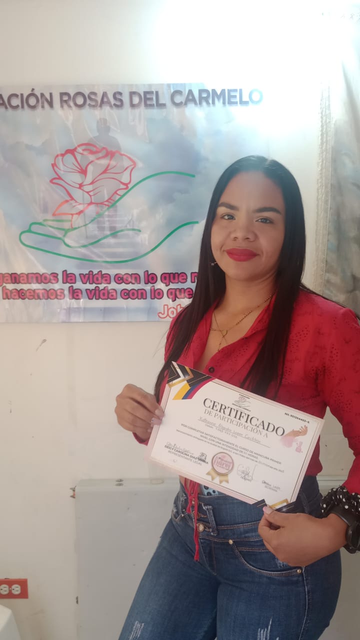 Graduada del curso de manicura sosteniendo su certificado frente al banner de la Fundación Rosas del Carmelo