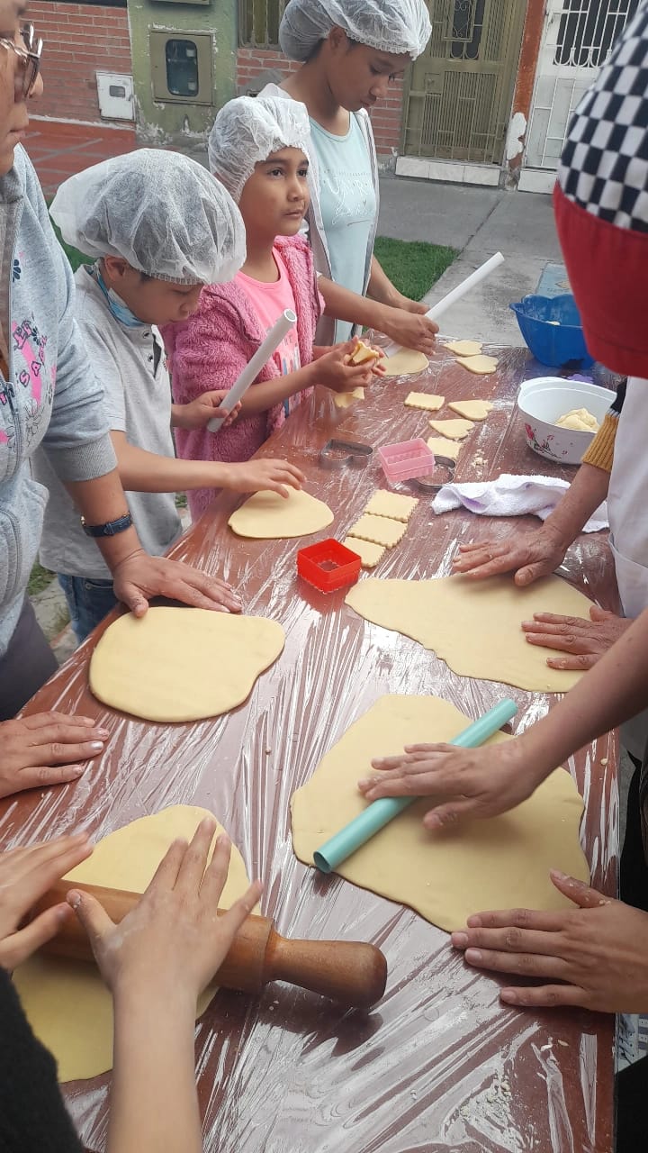Niños y adultos estirando y cortando figuras de masa sobre mesa larga en el curso de pastelería