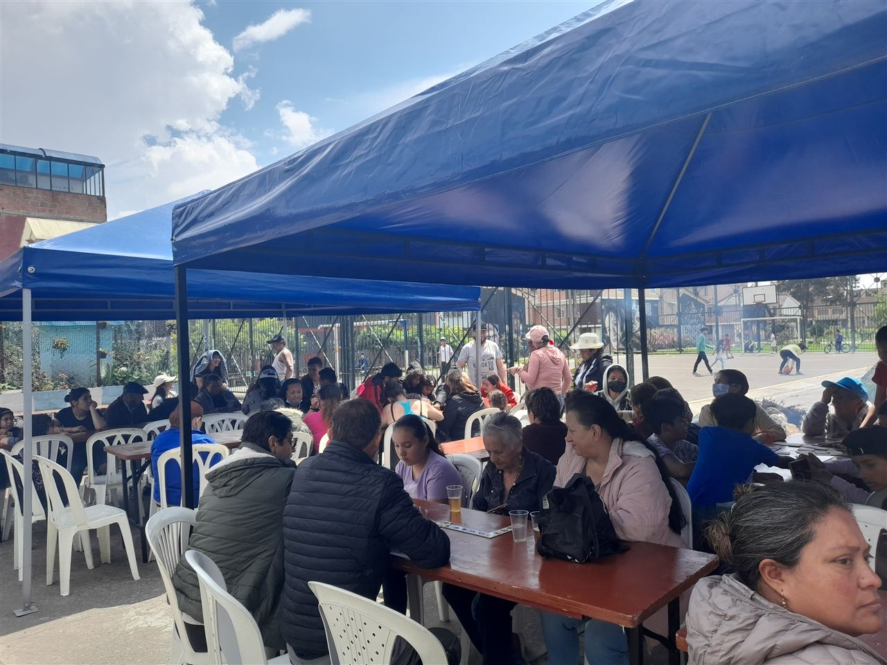 Familias sentadas bajo carpas azules en mesas comunitarias durante el Sancocho Comunitario, Fundación Rosas del Carmelo, Bogotá