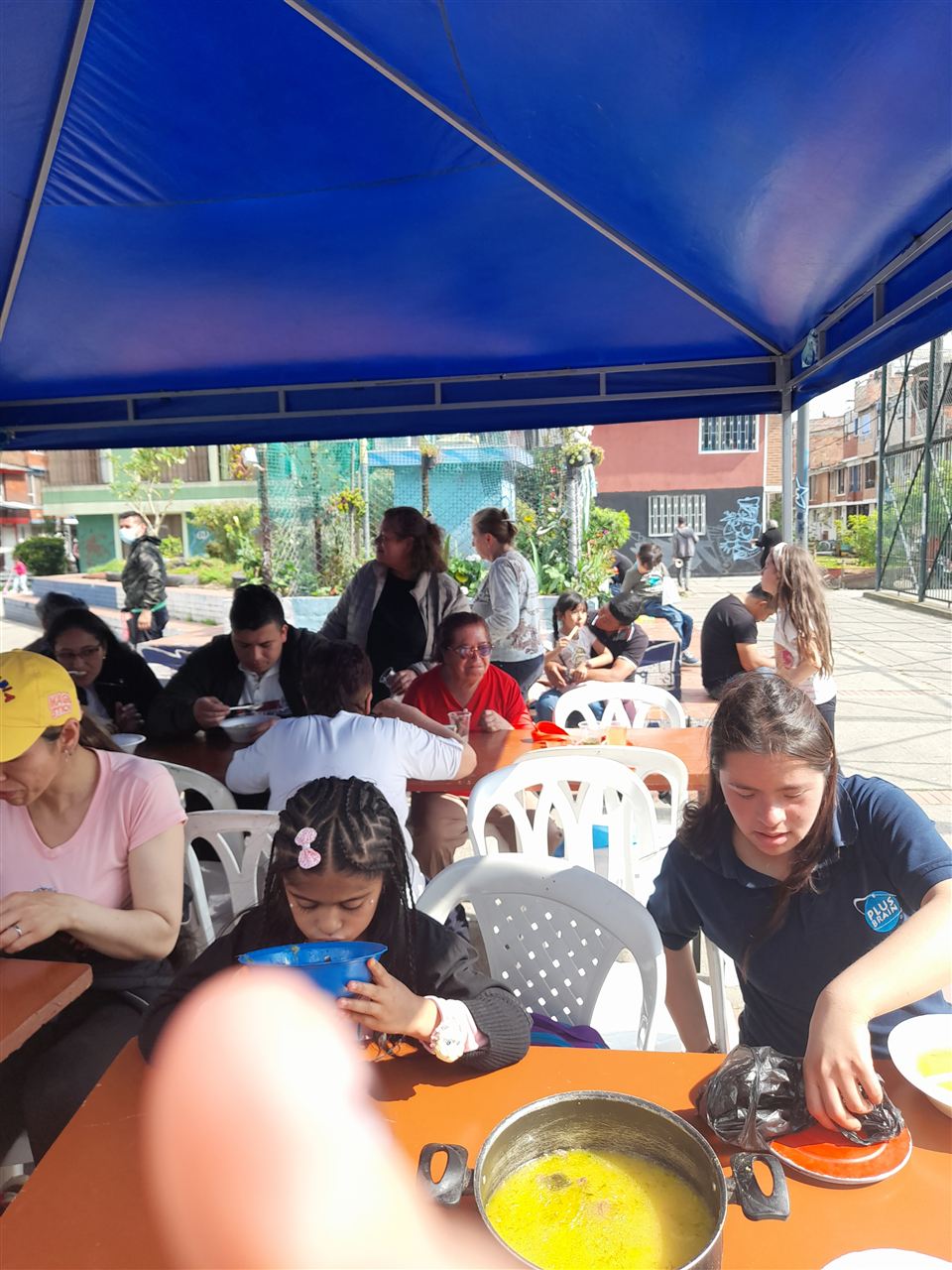 Niña comiendo sancocho en un tazón bajo una carpa durante la jornada comunitaria, Fundación Rosas del Carmelo, Bogotá